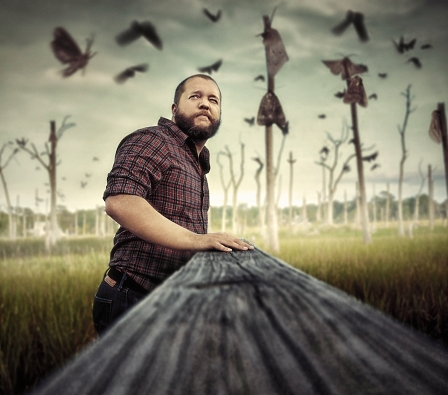 Der Musiker Radical Face steht an einem Holzbalken und trägt ein Hemd und Jeans 